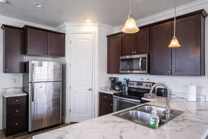 Kitchen featuring stainless steel appliances, dark wood finish cabinetry, light stone countertops, a textured ceiling, and hanging light fixtures