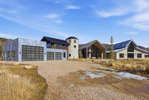 Gravel driveway with a multi-structure property featuring a modern garage building with three glass-paneled doors, a stone-clad silo-style tower, and two additional structures with contemporary pitched roofs and expansive window walls