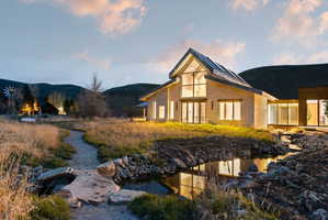 Stone exterior home featuring expansive glass panels, a steep gabled roofline, a decorative pond, and a winding gravel path through natural landscaping