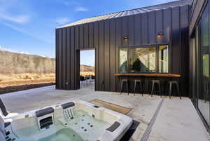 Outdoor patio featuring a built-in hot tub, concrete surfacing, a window bar with a wood-finish countertop, black exterior siding, and an open entry point to an interior space