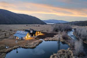 Modern-style residence featuring a dark facade with expansive windows, an attached light-colored structure with a metal roof, a pond, and a river winding through the landscape