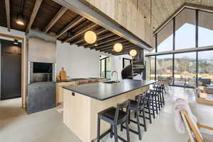 Kitchen featuring an island with sink, a breakfast bar area, concrete flooring, light wood finish cabinets, and a high wood beamed ceiling