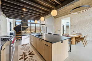 Open-concept living area featuring a large kitchen island with a dark countertop and integrated sink, a spiral staircase with wood treads, floor-to-ceiling windows, a stone accent wall, and exposed wood ceiling beams