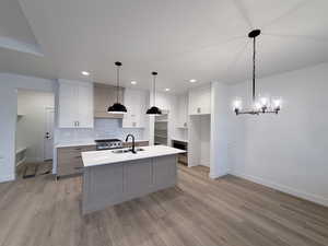 Two tone kitchen with two tone cabinetry, an island with sink, stainless steel gas stove, a chandelier, and decorative backsplash