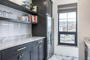 Kitchen with light stone counters, open shelves, freestanding refrigerator, and gray cabinets