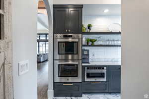Kitchen featuring stainless steel appliances, open shelves, light stone countertops, arched walkways, and gray cabinetry
