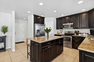 Kitchen featuring stainless steel appliances, dark wood finish cabinetry, light tile patterned floors, light stone counters, and a kitchen island