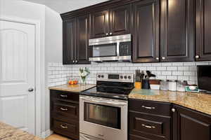 Kitchen with stainless steel appliances, dark wood finish cabinets, light stone countertops, and decorative backsplash