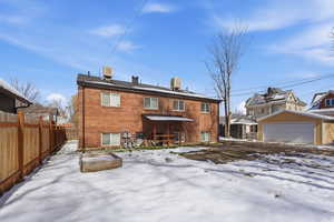 Snow covered house with a patio area, brick siding, a fenced backyard, and an outbuilding