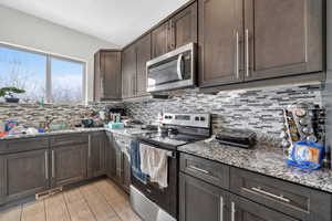 Kitchen with stainless steel appliances, dark wood finish cabinets, light stone countertops, light wood-style flooring, and backsplash