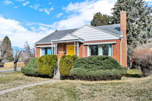 Bungalow-style home featuring a shingled roof, a front lawn, a chimney, brick siding, and covered porch