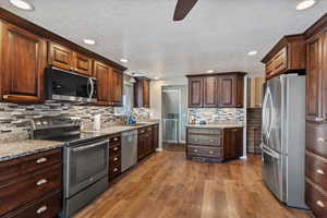 Kitchen with stainless steel appliances, dark wood-style flooring, dark wood finish cabinetry, a ceiling fan, and light stone counters