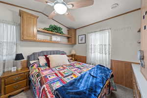 Carpeted bedroom featuring wood walls, a wainscoted wall, crown molding, a ceiling fan, and vaulted ceiling