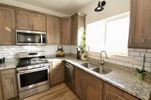 Kitchen featuring stainless steel appliances, backsplash, wood finish cabinets, light stone countertops, and light wood finished floors