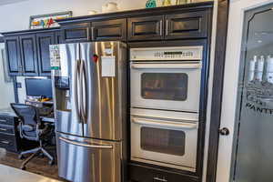 Kitchen featuring stainless steel refrigerator with ice dispenser, white double oven, a desk, dark wood-style floors, and dark cabinets