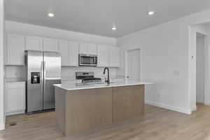 Two tone kitchen featuring stainless steel appliances, a kitchen island with sink, light wood finished floors, light stone countertops, and recessed lighting