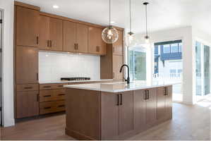 Kitchen with light wood-type flooring, a kitchen island with sink, decorative light fixtures, light stone countertops, and tasteful backsplash