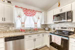 Kitchen with stainless steel appliances, white cabinets, vaulted ceiling, and light stone counters