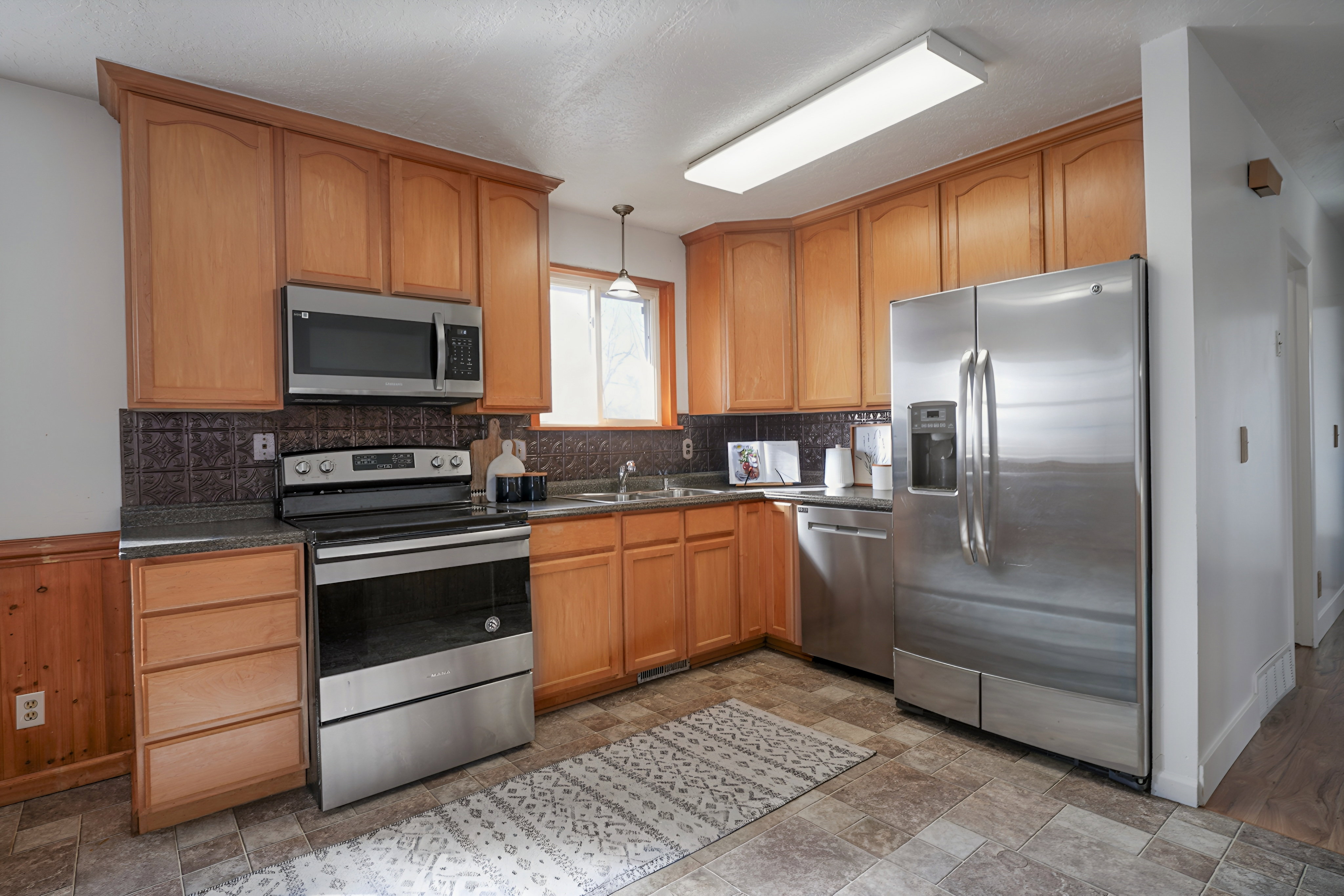 Kitchen with stainless steel appliances, pendant lighting, dark countertops, stone finish flooring, and wood finish cabinetry