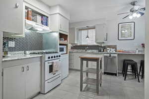 Kitchen featuring white appliances, white cabinetry, backsplash, a ceiling fan, and light stone counters