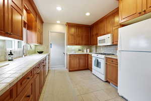 Kitchen featuring white appliances, light tile patterned floors, wood finish cabinets, tile counters, and recessed lighting
