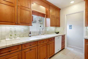 Kitchen featuring wood finish cabinetry, dishwasher, tile counters, light tile patterned floors, and decorative backsplash