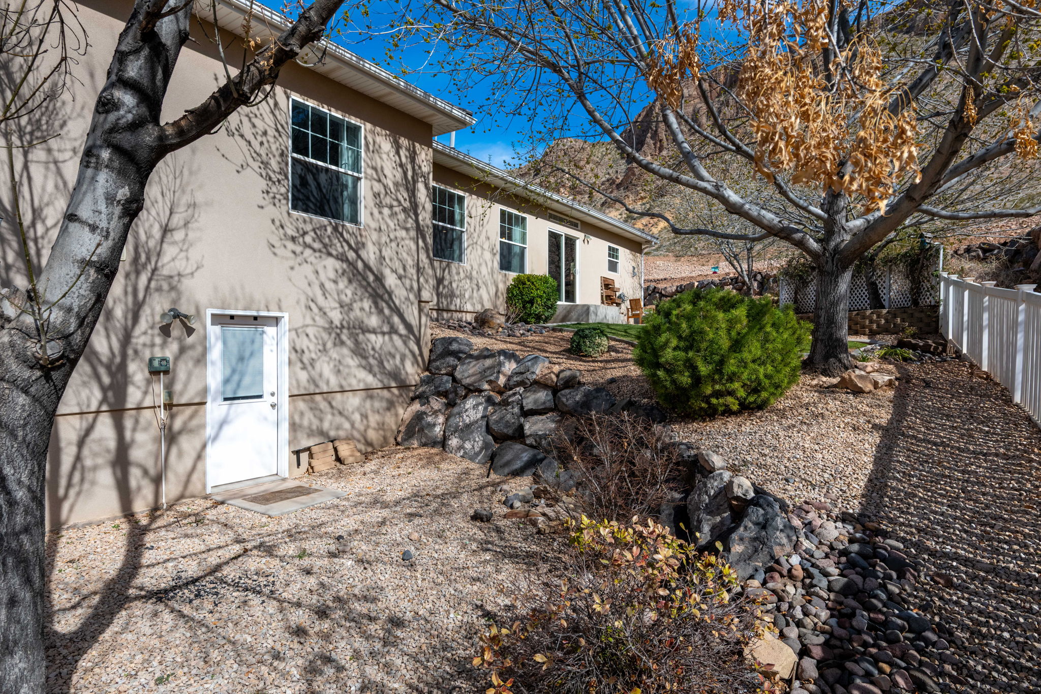 Terraced landscaping featuring a white picket fence, a mature deciduous tree, a large evergreen shrub, decorative rock beds, and a stone retaining wall