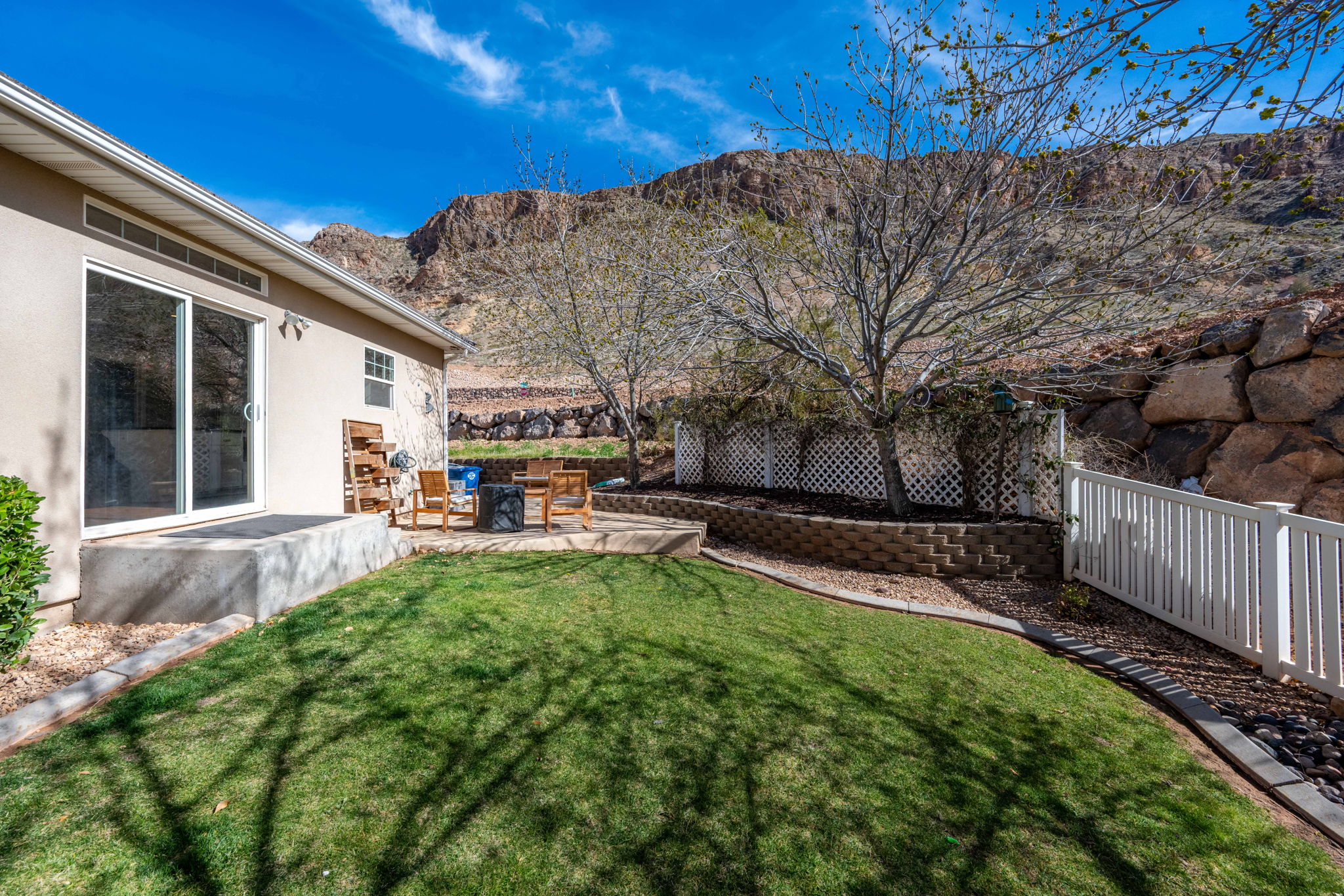 Sliding glass door access to a patio with stone pavers, bordered by a retaining wall, a white lattice fence, and a white picket fence