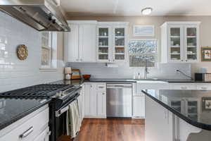 Kitchen featuring range with gas cooktop, dark stone counters, stainless steel dishwasher, white cabinets, and glass fronted cabinets