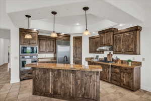 Kitchen with dark wood finish cabinets, a raised ceiling, decorative light fixtures, built in appliances, and a center island with sink