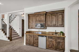 Indoor wet bar with light colored carpet, stainless steel appliances, dark wood finish cabinets, and tile counters