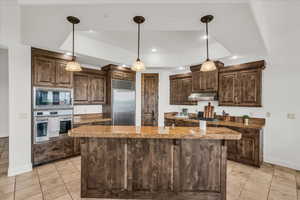 Kitchen with dark wood finish cabinets, built in appliances, pendant lighting, a raised ceiling, and a center island