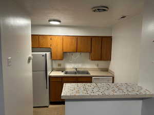 Kitchen with wood finish cabinets, freestanding refrigerator, light countertops, white dishwasher, and a textured ceiling