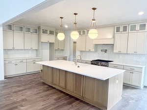 Kitchen featuring glass insert cabinets, a center island with sink, backsplash, pendant lighting, and light stone counters