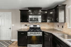 Kitchen with dark cabinets, stainless steel appliances, light stone countertops, and backsplash
