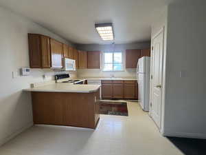 Kitchen featuring a peninsula, wood finish cabinets, light countertops, white appliances, and light flooring