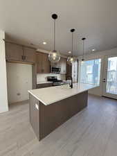 Kitchen featuring light wood-style flooring, stainless steel appliances, pendant lighting, a center island with sink, and light stone counters