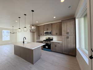 Kitchen featuring stainless steel appliances, an island with sink, light wood-style flooring, and decorative light fixtures