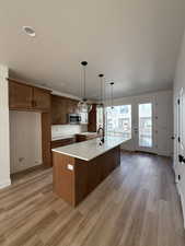 Kitchen with light stone countertops, a center island with sink, light wood-style flooring, wood finish cabinetry, and decorative light fixtures