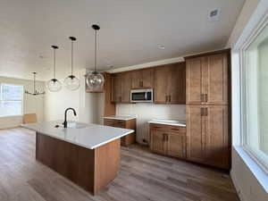 Kitchen with wood finish cabinetry, a kitchen island with sink, hanging light fixtures, stainless steel microwave, and light wood-style flooring