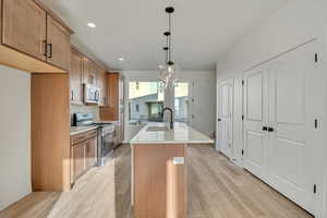 Kitchen with a kitchen island with sink, stainless steel appliances, wood finish cabinetry, light wood-style floors, and light stone counters