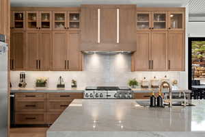 Kitchen with glass fronted cabinets, wood finish cabinetry, light stone counters, decorative backsplash, and stainless steel gas range