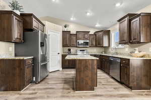 Kitchen with dark wood finish cabinets, stainless steel appliances, a kitchen island, light stone countertops, and light wood-type flooring