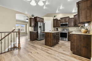 Kitchen with dark wood finish cabinets, stainless steel appliances, a kitchen island, lofted ceiling, and light stone counters