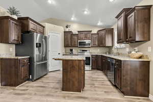 Kitchen featuring dark wood finish cabinetry, stainless steel appliances, vaulted ceiling, light stone counters, and light wood-style flooring