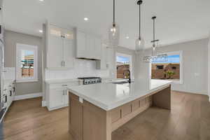 Two tone kitchen with glass fronted cabinets, pendant lighting, light wood-style floors, an island with sink, and light stone counters
