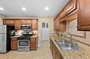 Kitchen featuring stainless steel appliances, wood finish cabinetry, light stone counters, light tile patterned floors, and tasteful backsplash