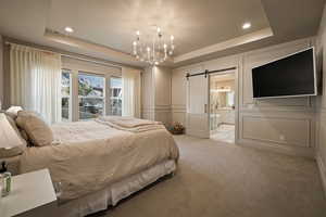 Carpeted bedroom with a barn door, a decorative wall, a chandelier, a tray ceiling, and connected bathroom