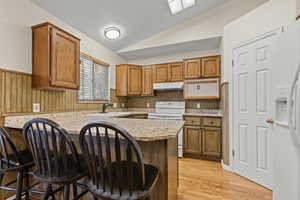 Kitchen featuring a wainscoted wall, a peninsula, wood finish cabinets, white appliances, and vaulted ceiling