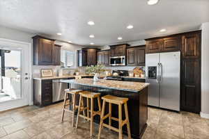 Kitchen with stainless steel appliances, dark wood finish cabinets, large center island, light stone countertops, and recessed lighting